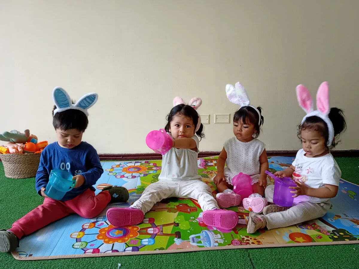 Cuatro niños con orejas de conejito sentados en tapete de juego — celebración de Pascua en guardería Brisa de Hogar